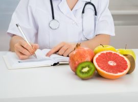 Close-up Of Female Dietician Writing Prescription With Fruits On Desk